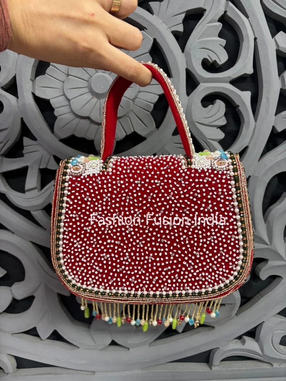 Red handbag with white polka dots and colorful tassels held by a hand against an ornate gray background.