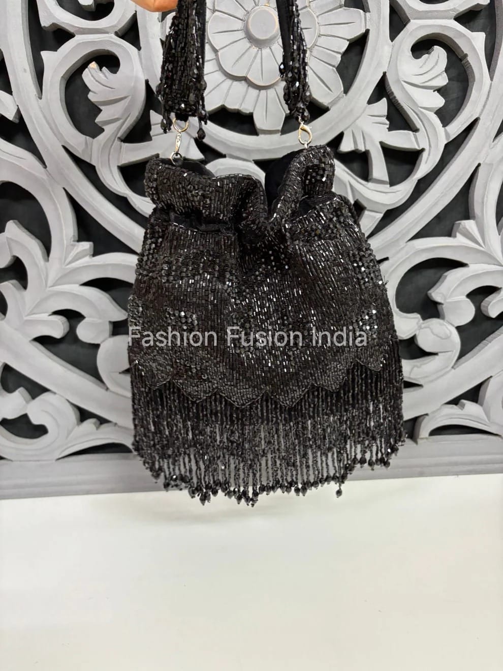 Black sequin handbag with fringe details against a decorative white and gray background.
