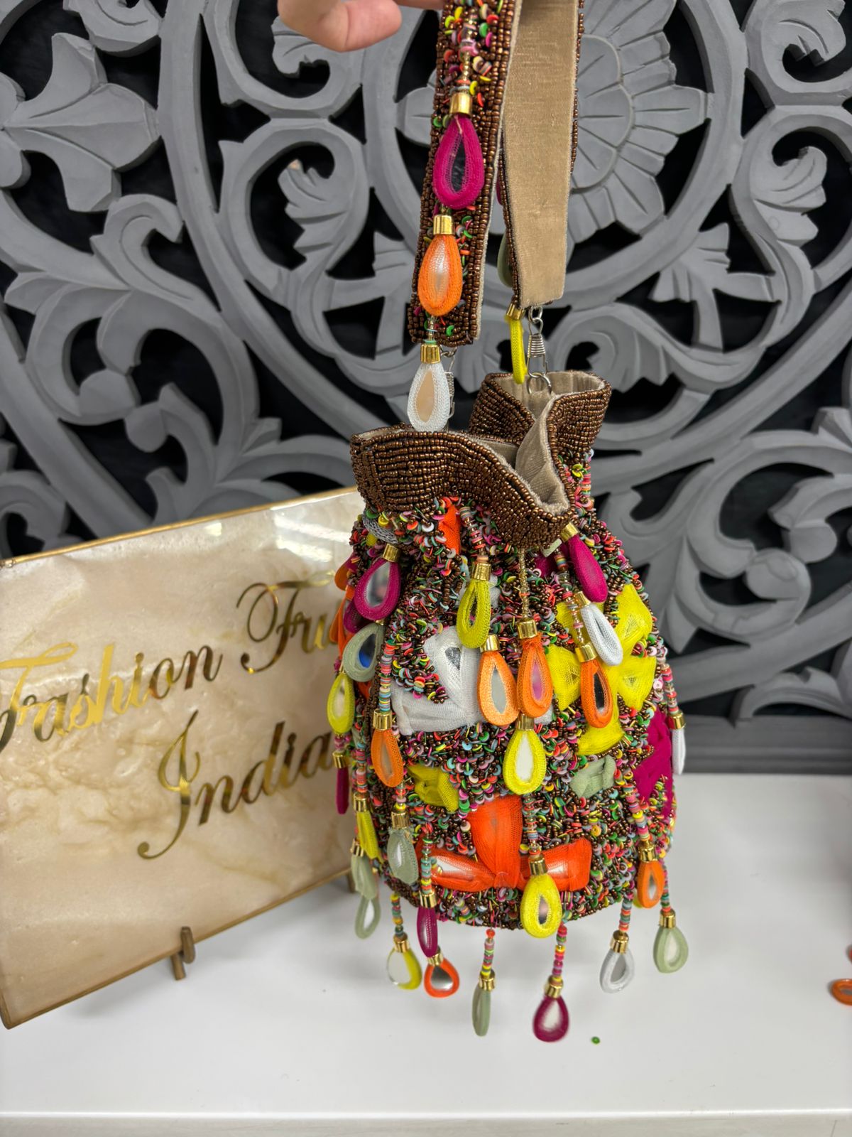 Decorative hanging ornament with colorful beads and stones on a white surface with 'Fashion From India' sign.