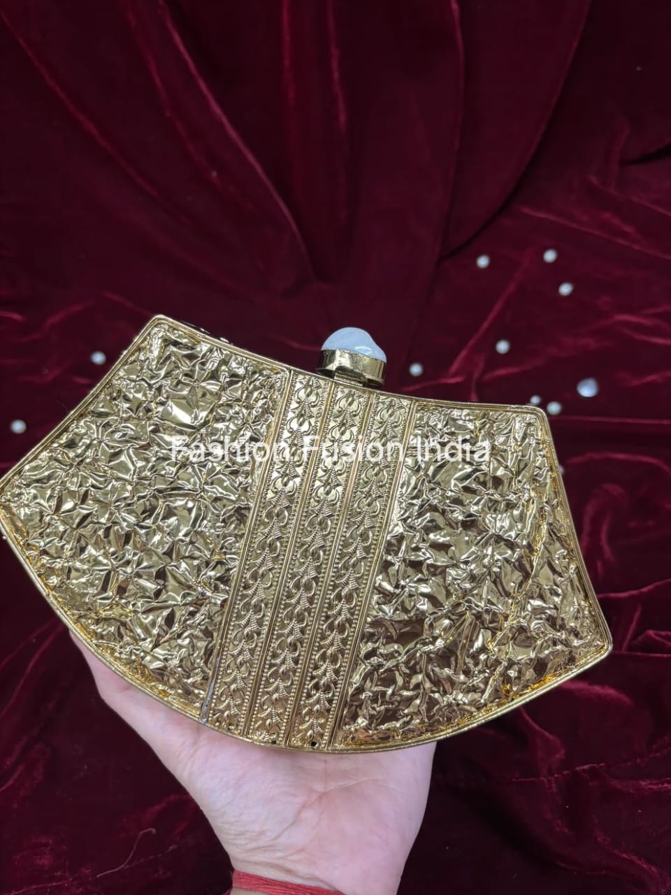 Gold clutch bag with intricate designs held by a hand against a red fabric background.