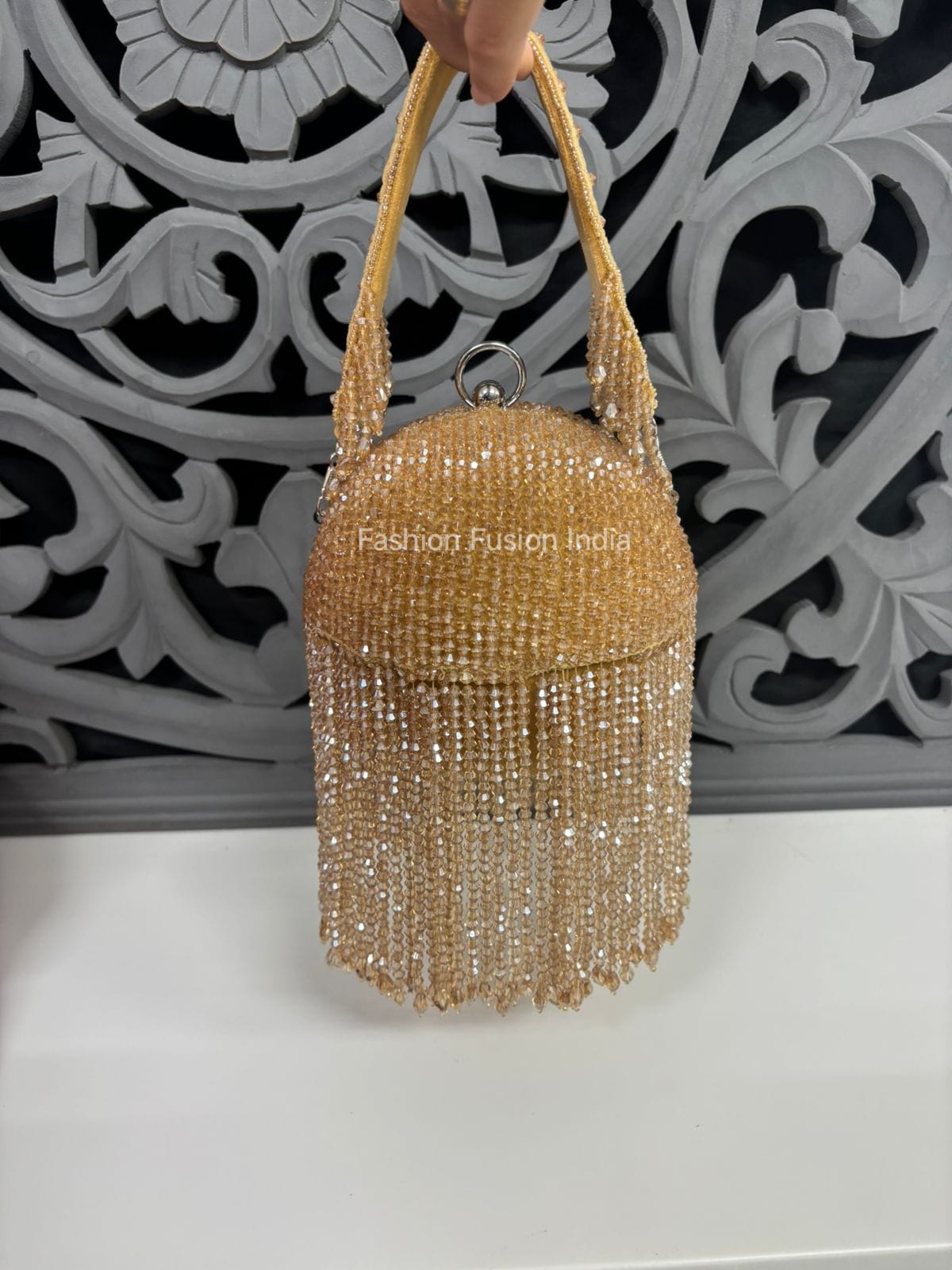 Gold beaded handbag held by a hand against an ornate white and gray decorative wall.