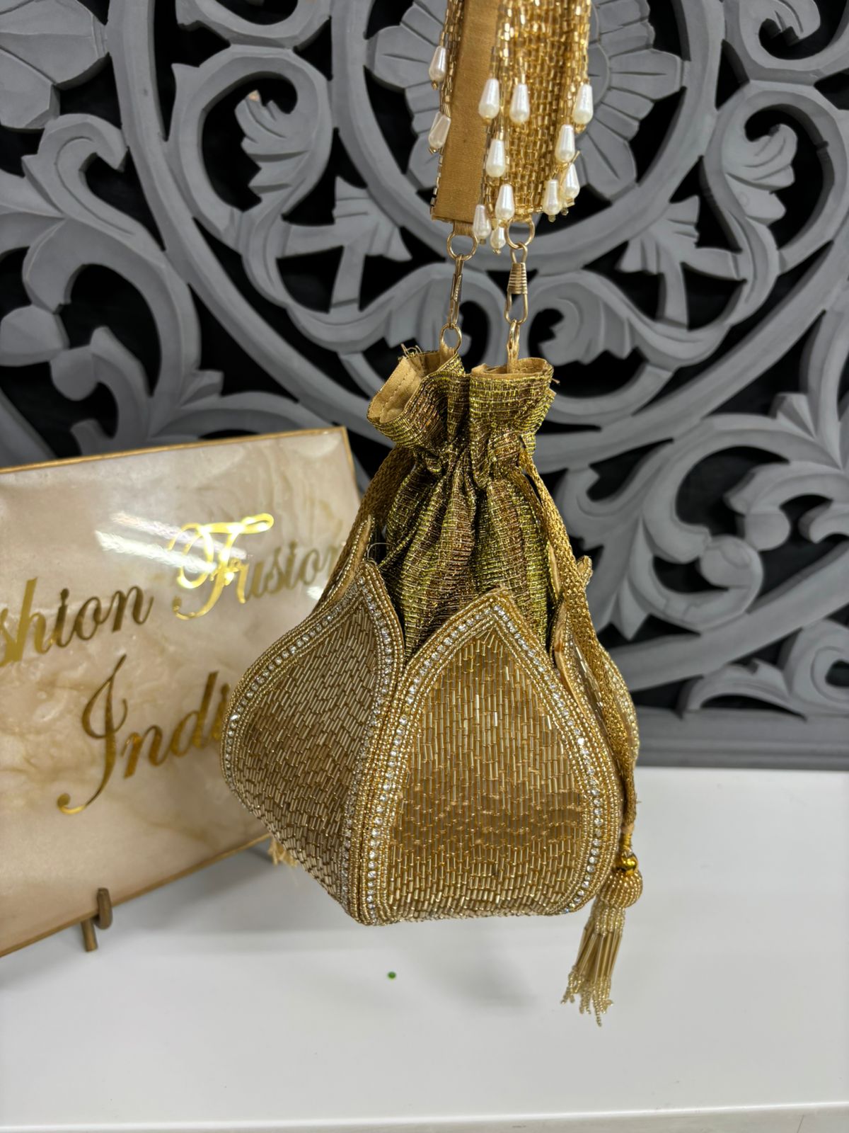 Gold beaded handbag with decorative elements on a white surface with a patterned wall in the background.