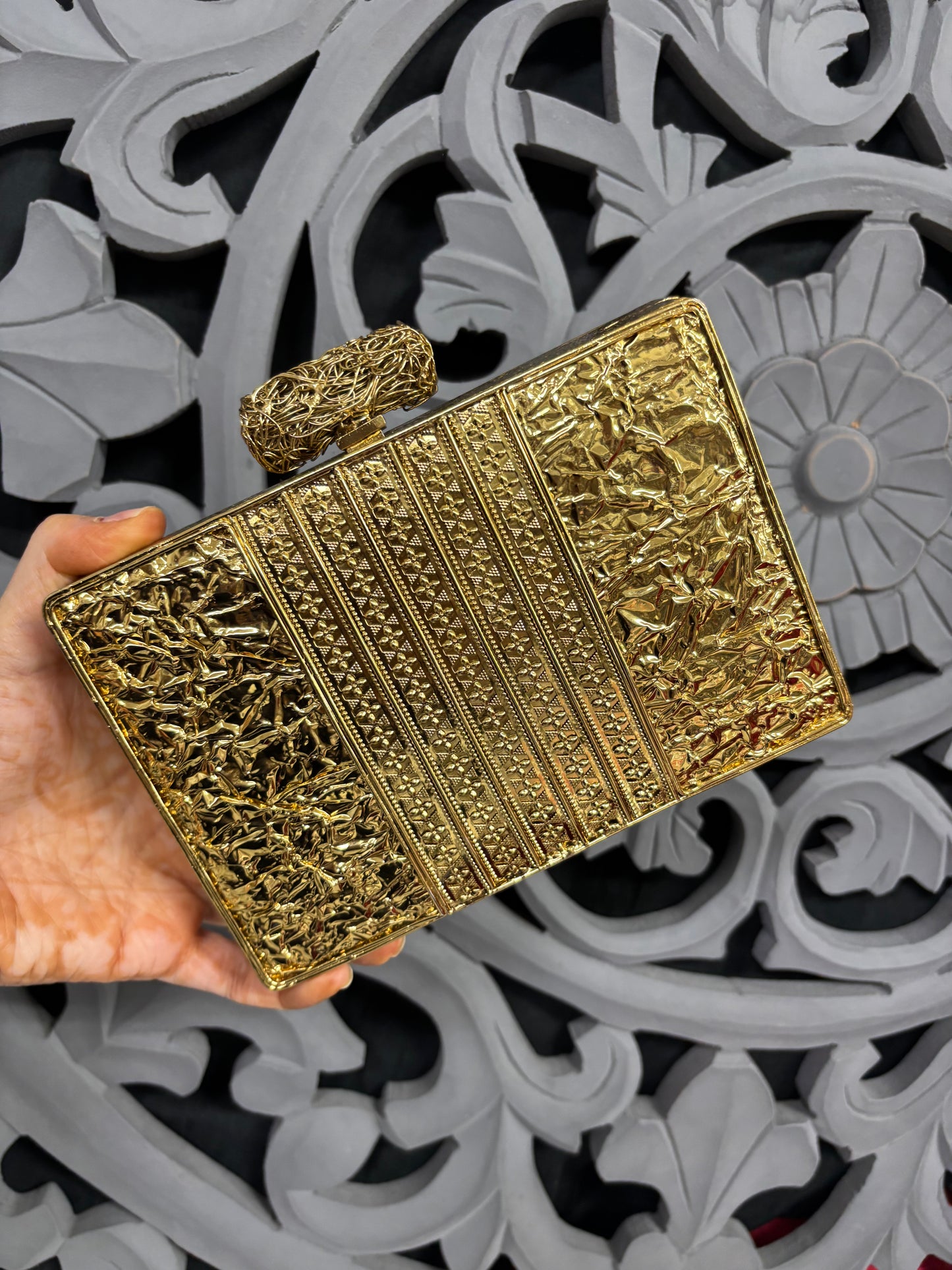 Gold clutch held by a hand against an ornate, patterned background