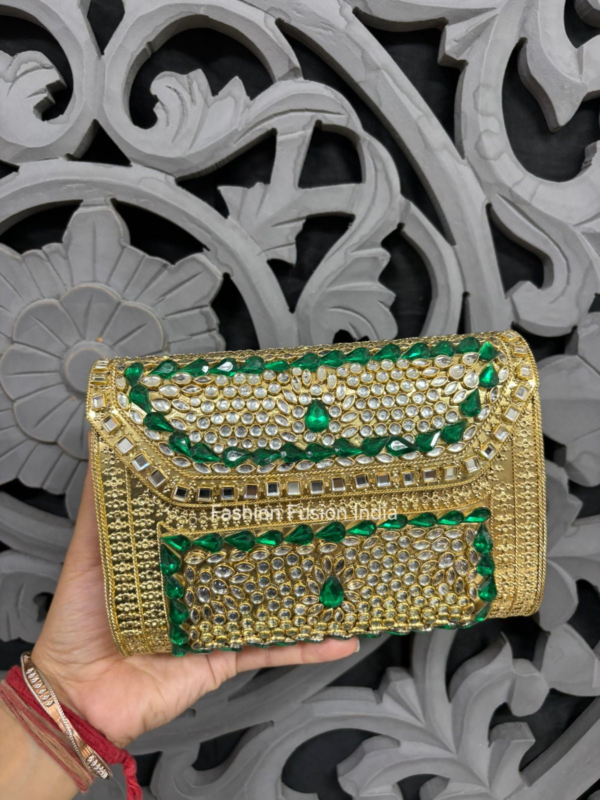 Gold and green embellished clutch held by a hand against an ornate gray background