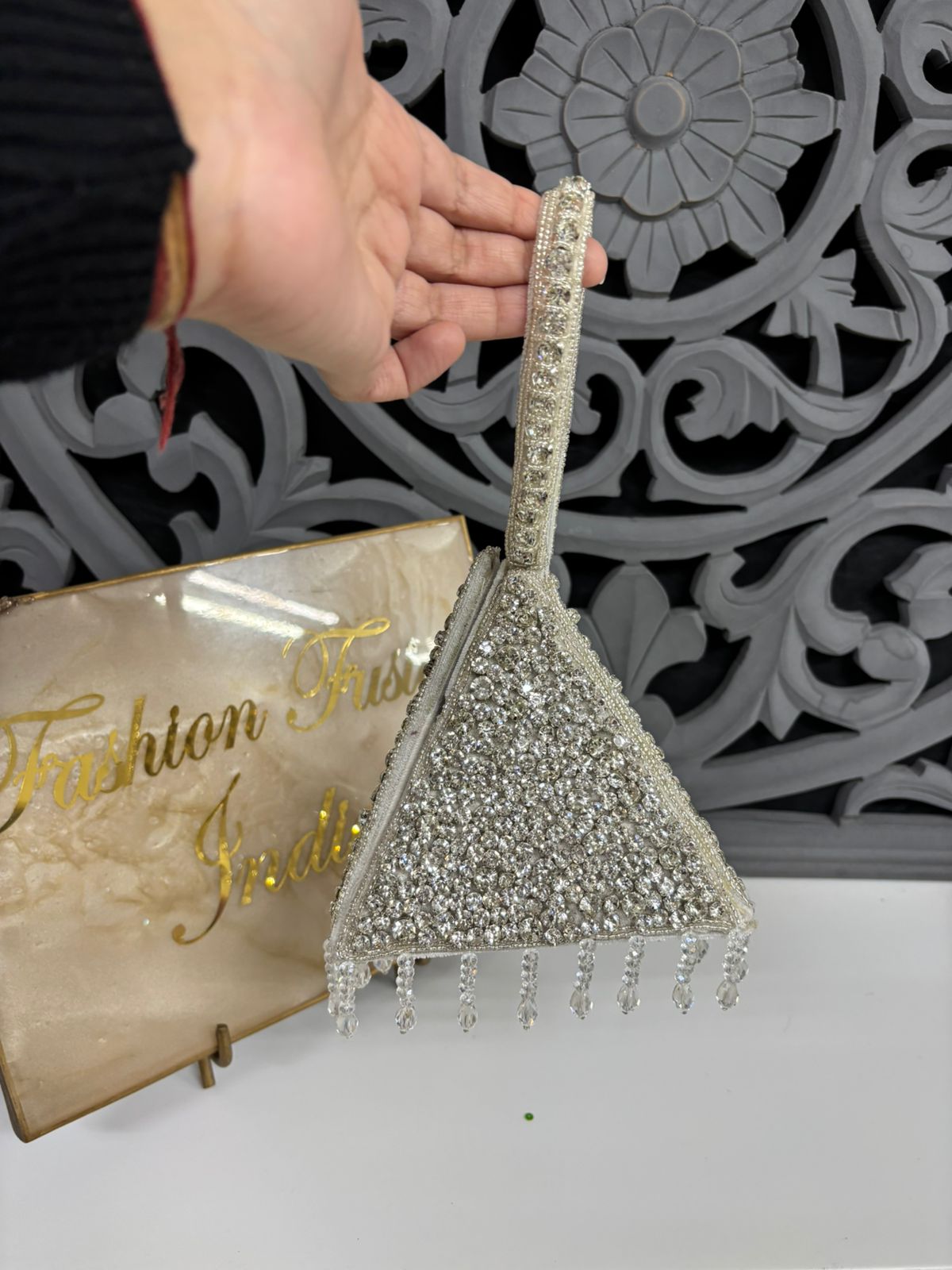 Hand holding a decorative silver clutch with intricate design against a patterned wall.