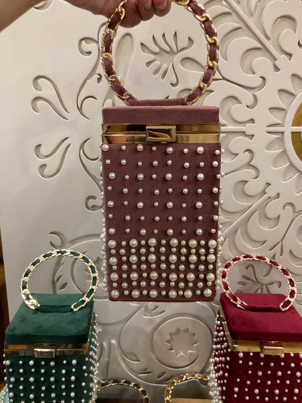 Mauve Pearl handbag with pearl embellishments held by a hand against an ornate white background.