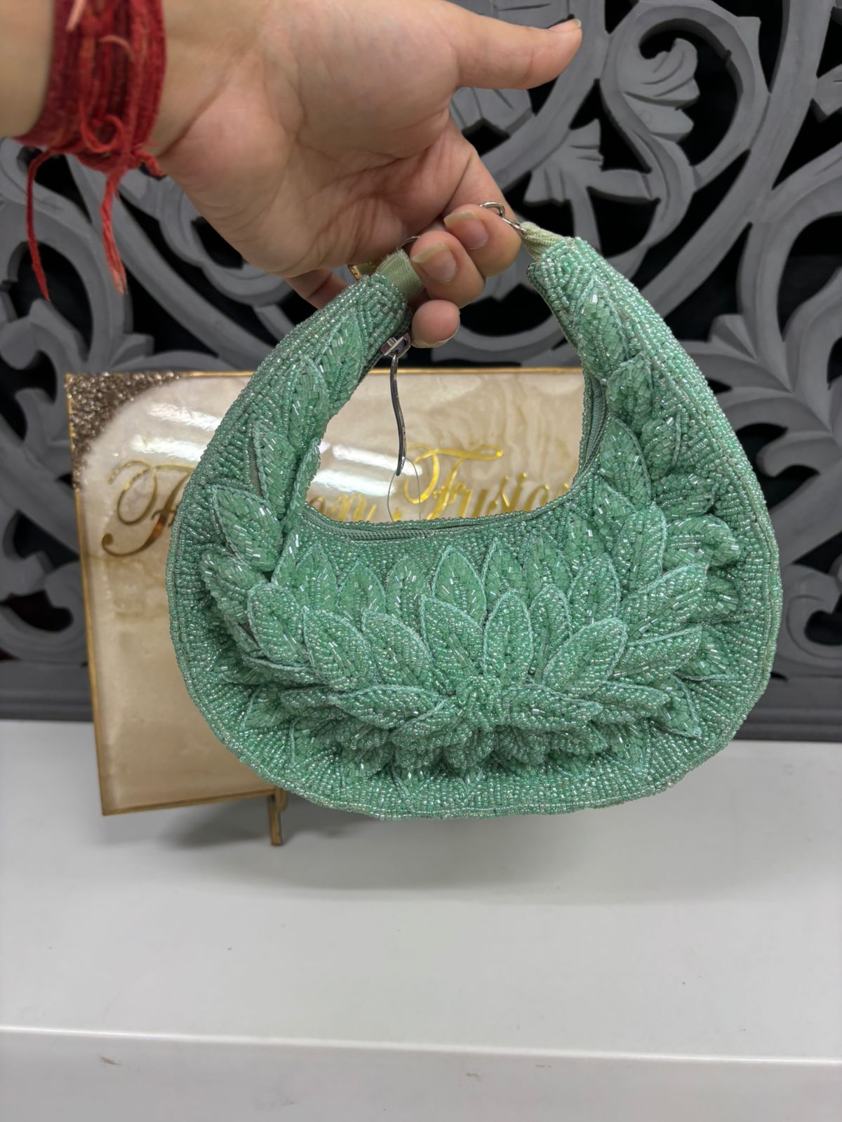 Green textured handbag held by a hand with a red bracelet against a decorative background
