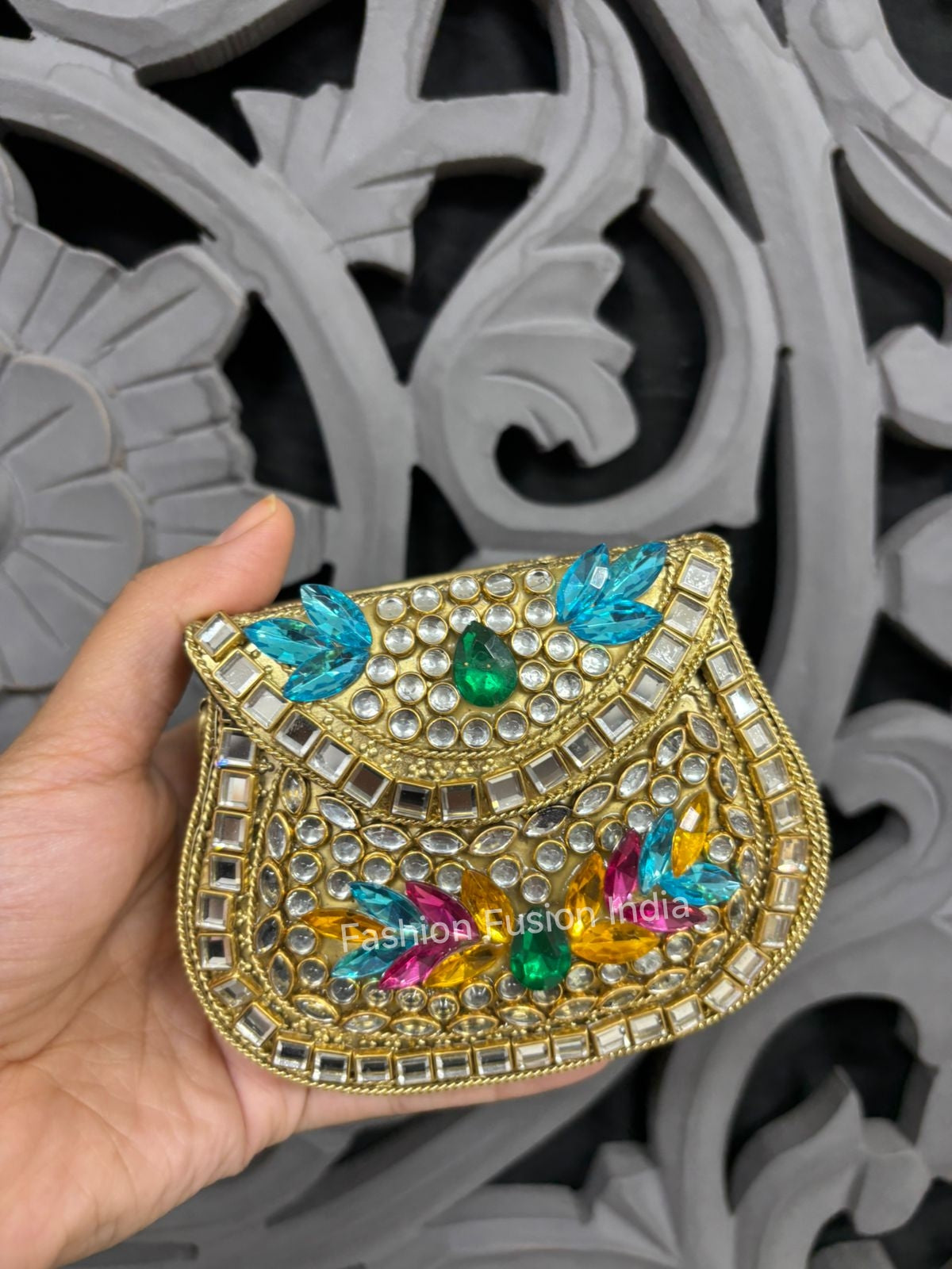 Decorative handbag with colorful gemstones held by a hand against a decorative gray background.
