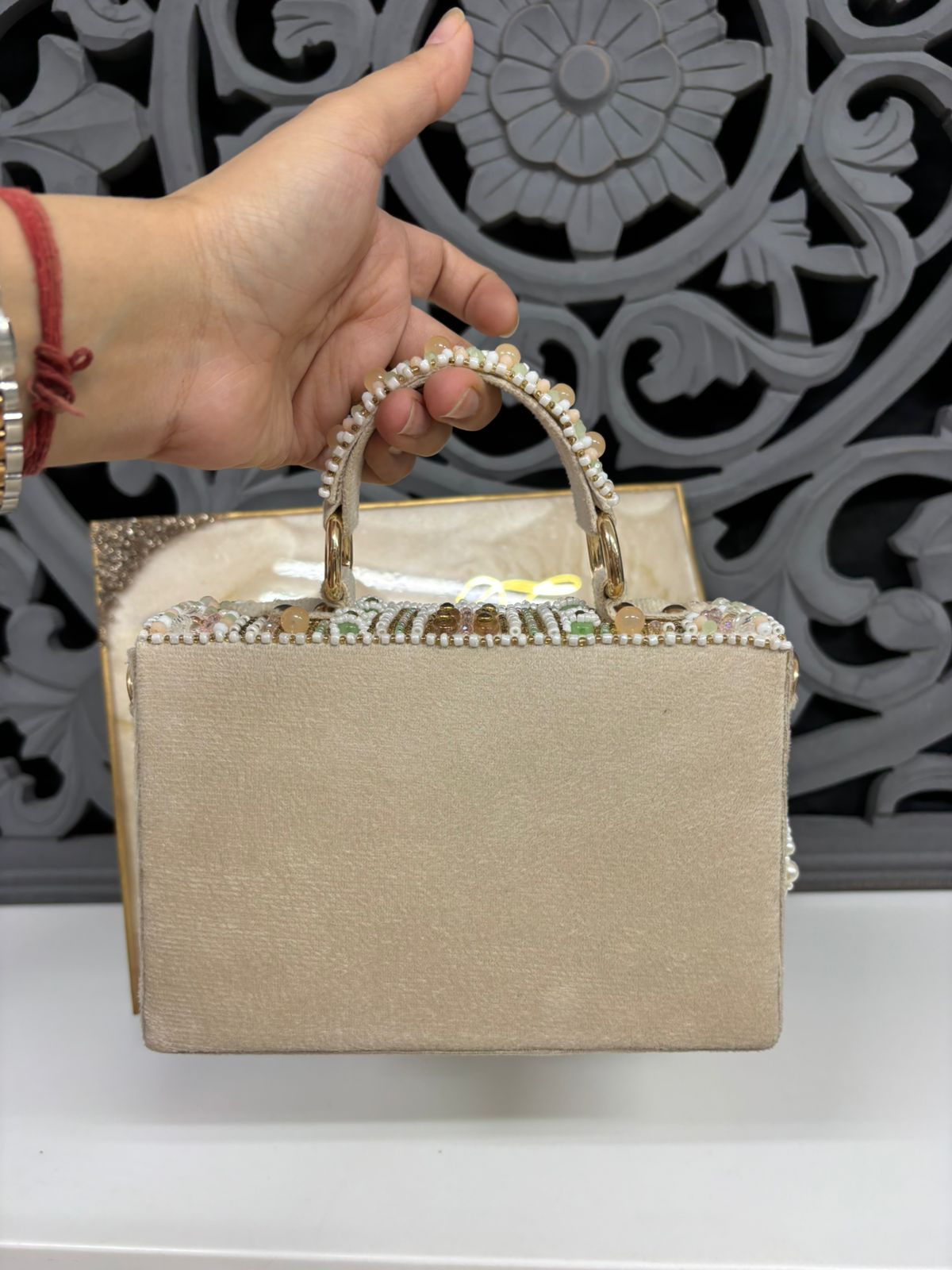 Beige handbag with decorative handle held by a hand against an ornate background