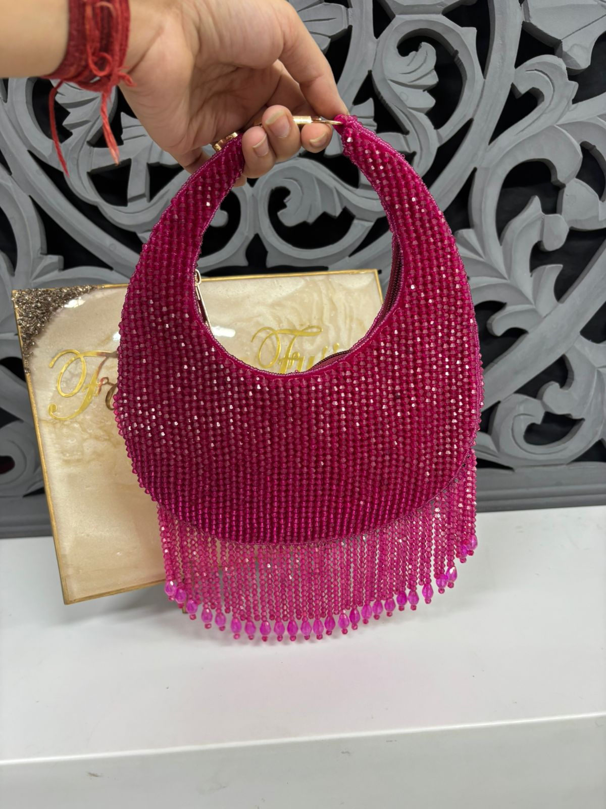 Pink beaded handbag held by a hand with a red bracelet against a decorative background.