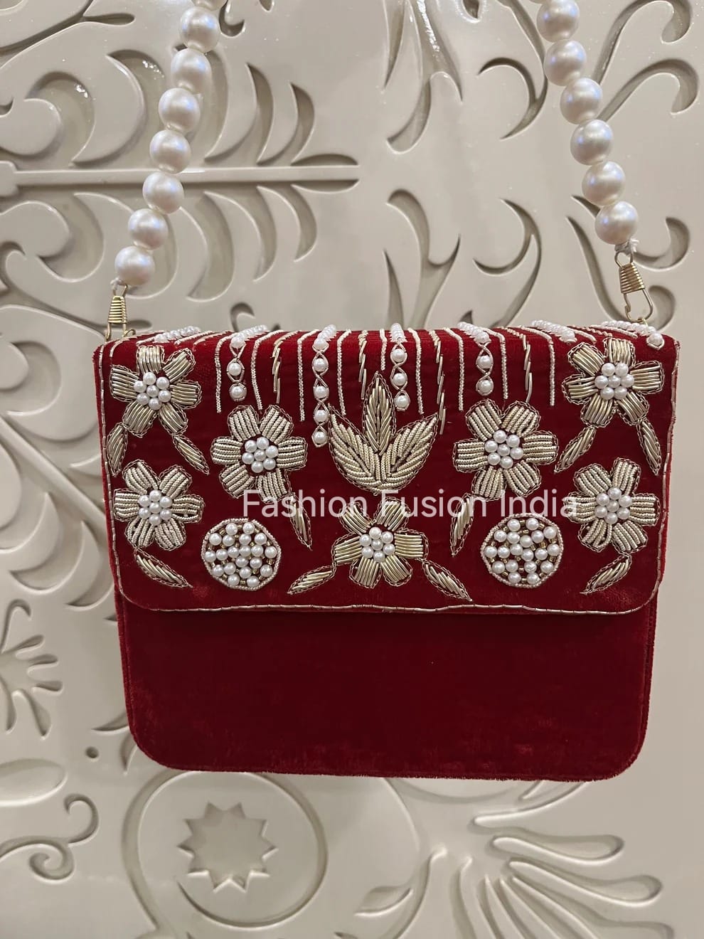 Red velvet clutch with floral embroidery and pearl details on a decorative white background.