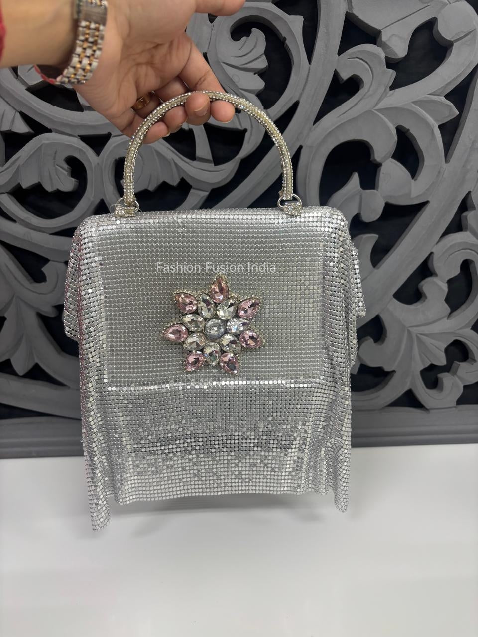 Silver handbag with decorative elements held by a hand against an ornate wall.