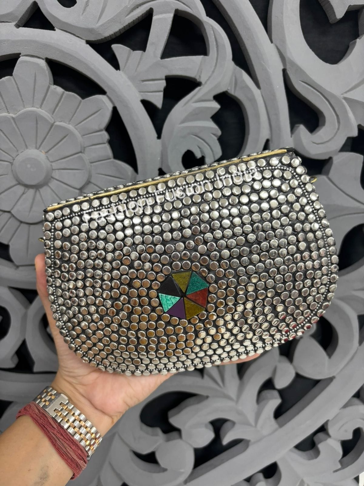 Silver studded clutch with a colorful center held by a hand against a decorative gray background