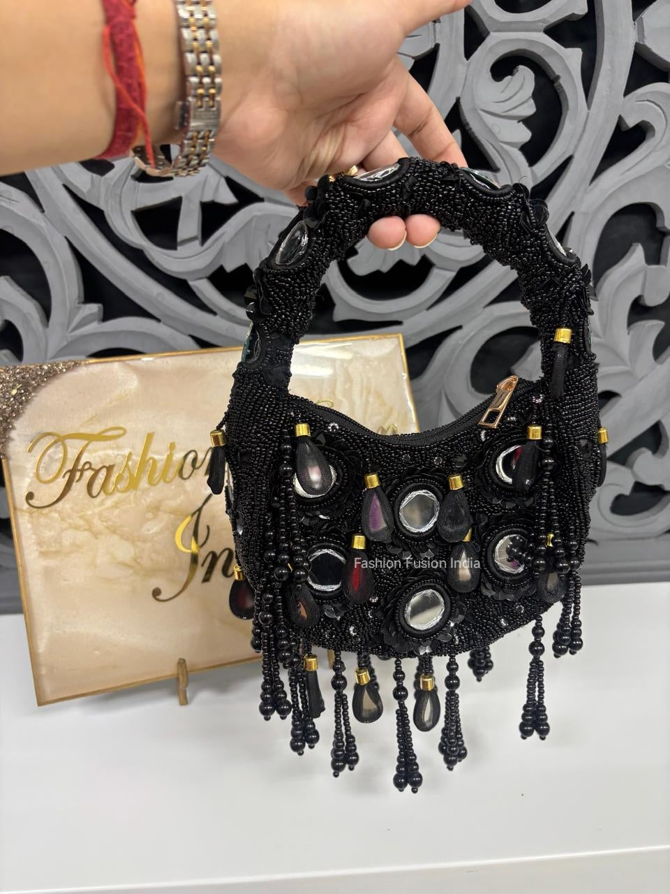 Black beaded handbag with tassels held by a hand against a decorative background.