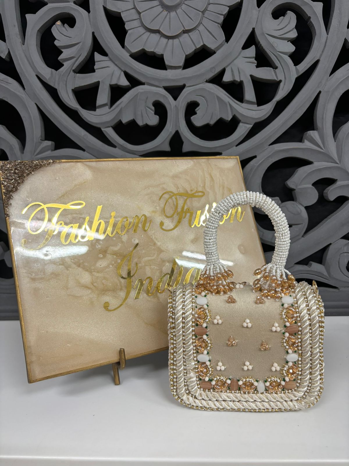 Decorative handbag with floral patterns on a stand, with 'Fashion From India' sign in the background.