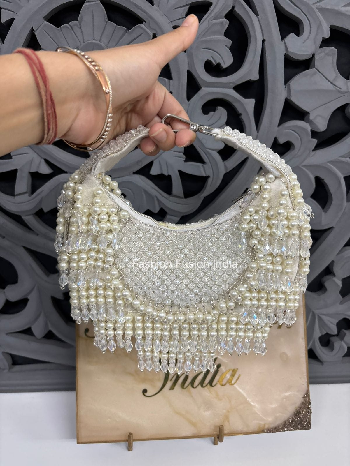 Hand holding a beaded handbag with 'Fashion Elegance India' branding against a decorative wall.