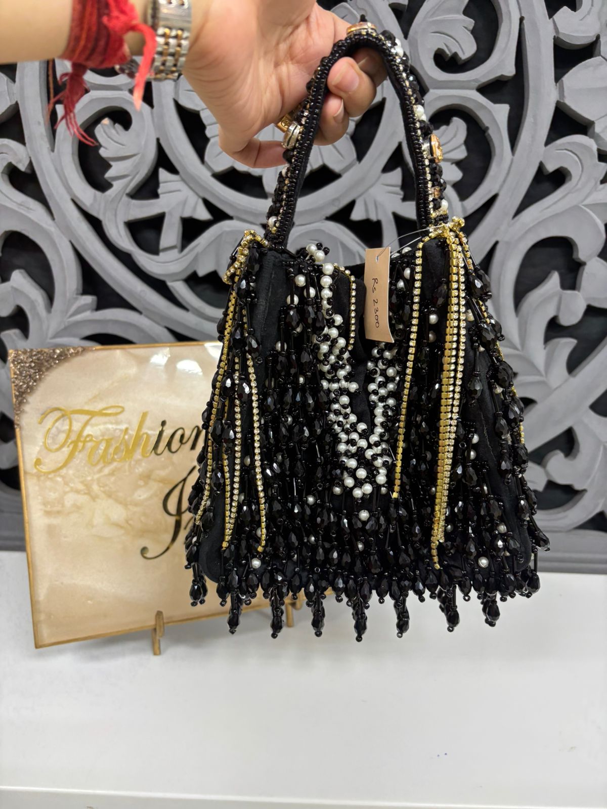 Black beaded handbag with tassels held by a hand against a decorative white and black background.