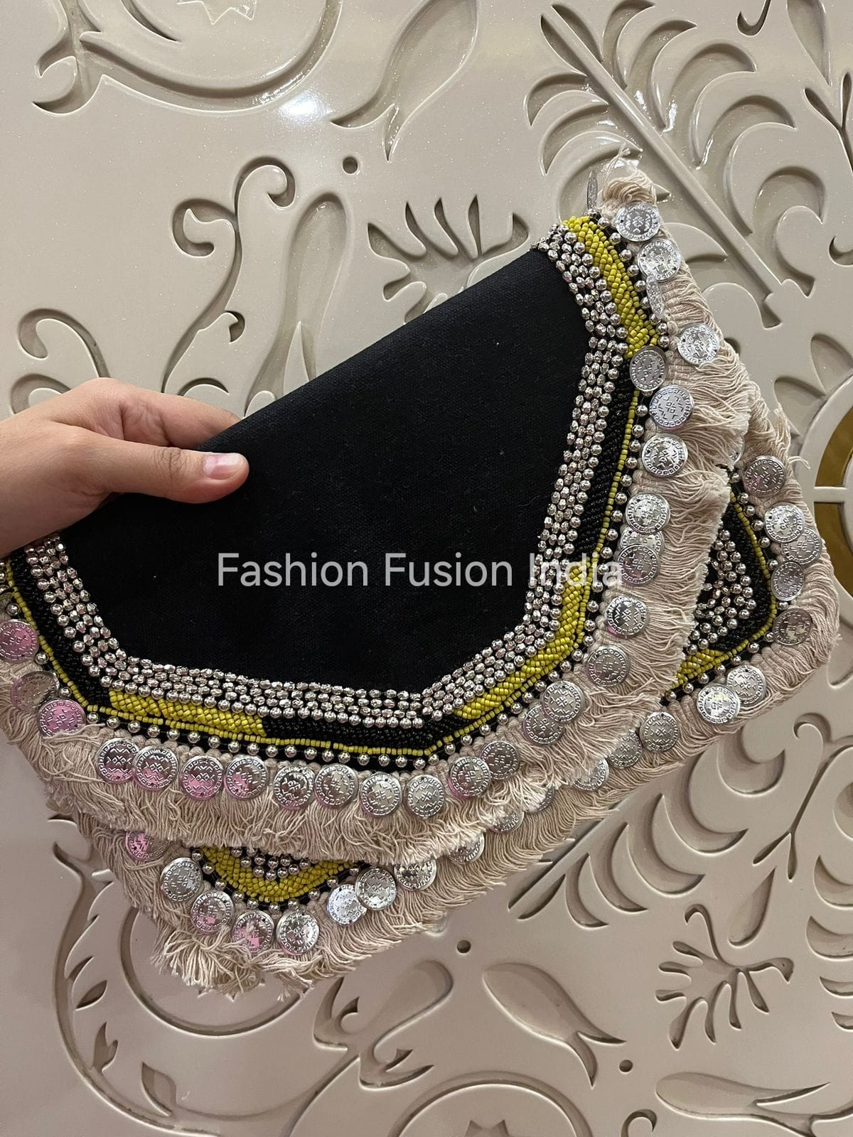 Black clutch with decorative embellishments held by a hand against an ornate background, with 'Fashion Fusion India' branding.