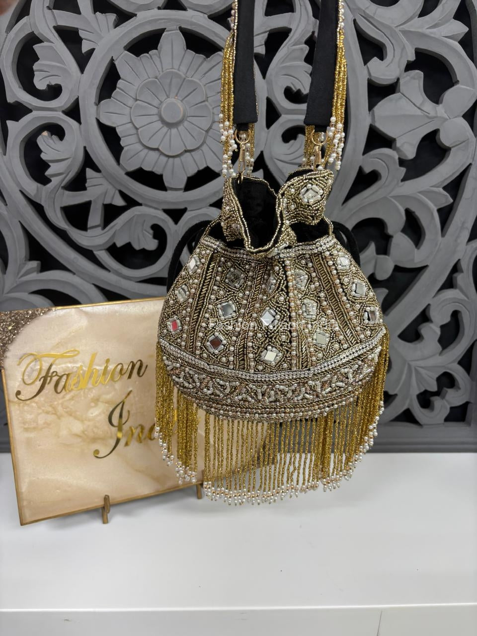 Decorative handbag with gold fringe and embellishments on a white surface with a decorative wall in the background.