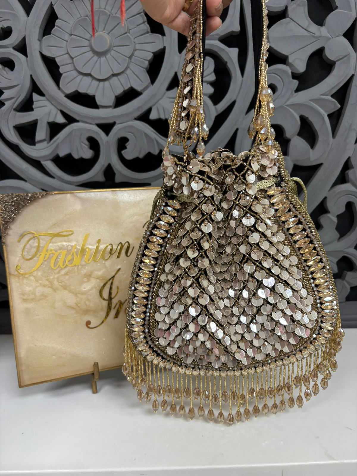 Decorative handbag with beads and tassels held by a hand, with 'Fashion India' sign in the background.