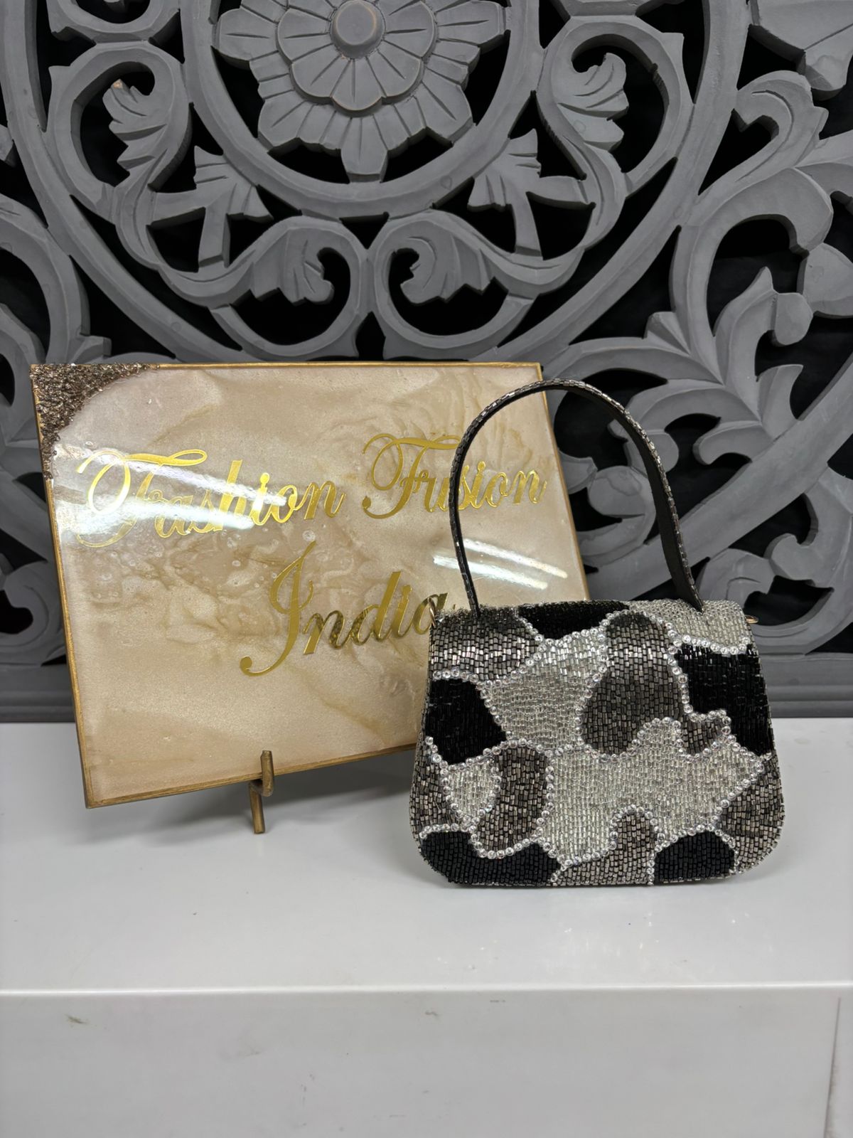 Black and silver floral patterned handbag with a gold card in front of a decorative wall.