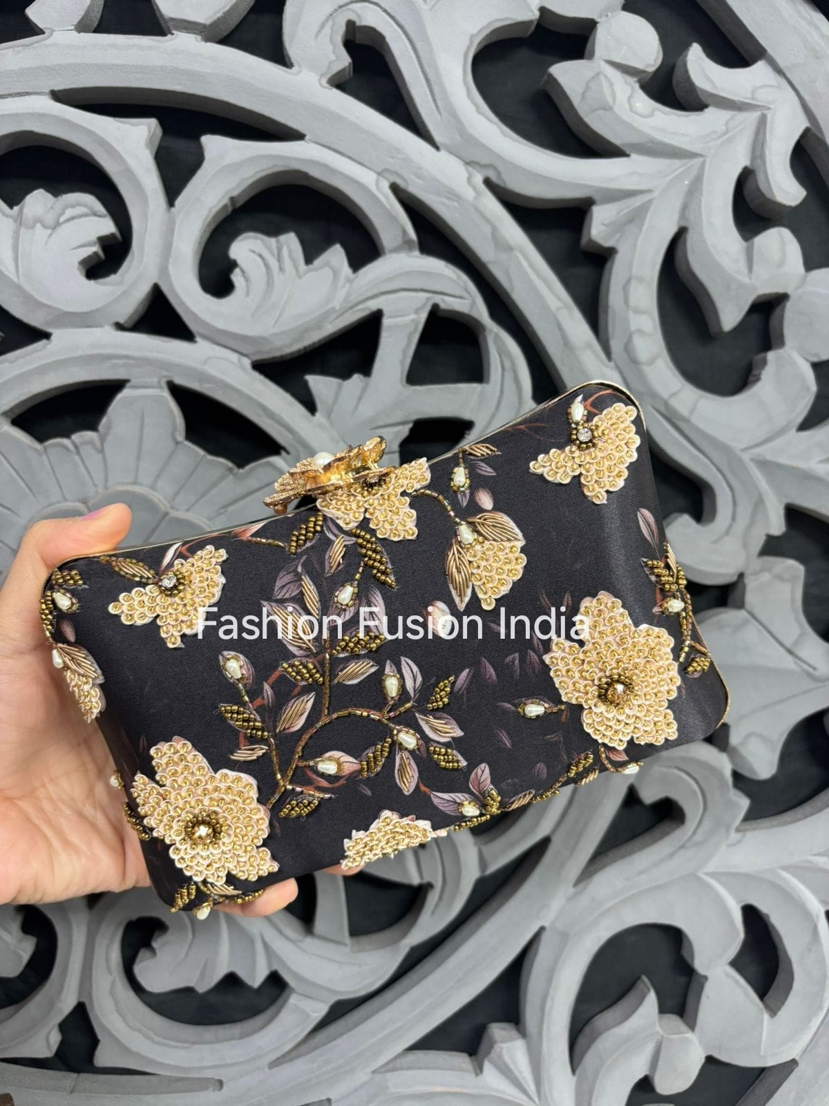 Black clutch with gold floral embroidery held by a hand against an ornate black and white background.