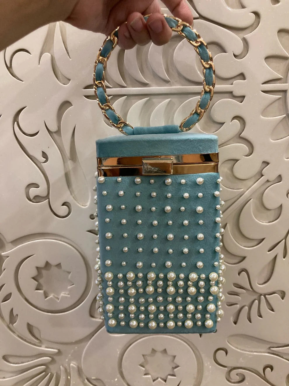 Blue handbag with pearl embellishments held by a hand against an ornate white background