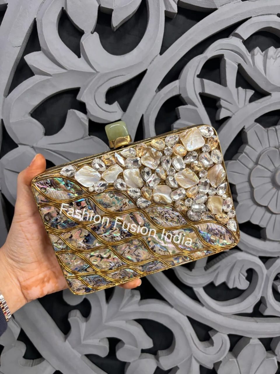 Gold clutch with stone embellishments held by a hand against an ornate gray background.