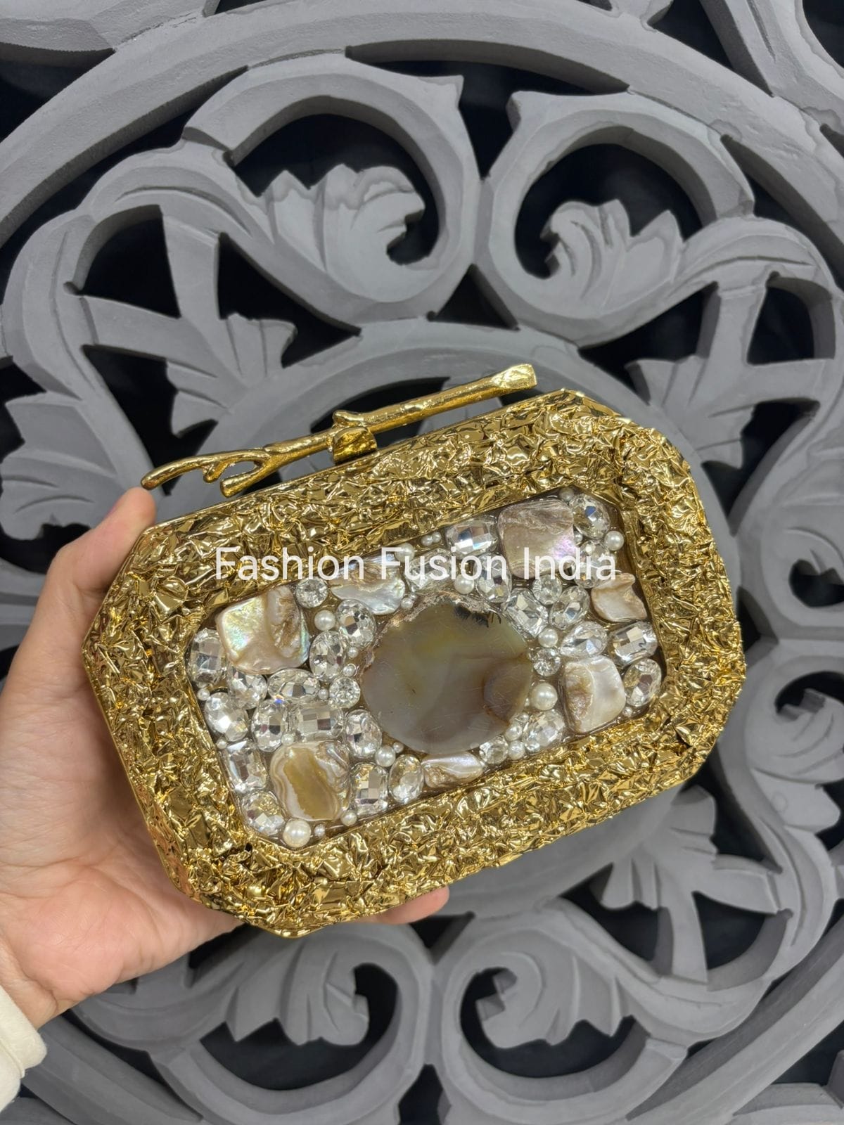 Gold clutch with gemstone embellishments held by a hand against a decorative gray background.