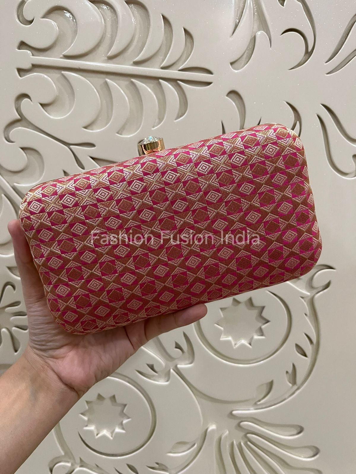 Pink clutch bag with gold patterns held by a hand against an ornate white background.