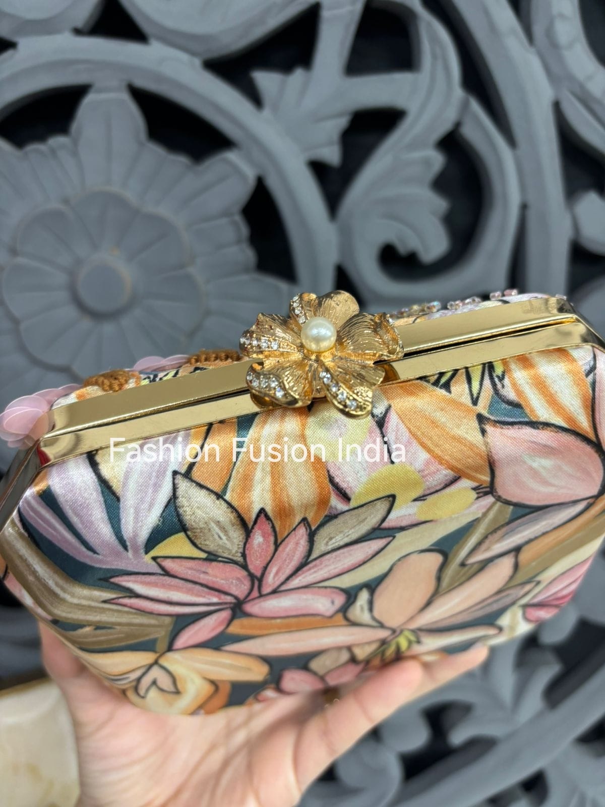 Floral patterned clutch with gold accents held by a hand against a decorative background.