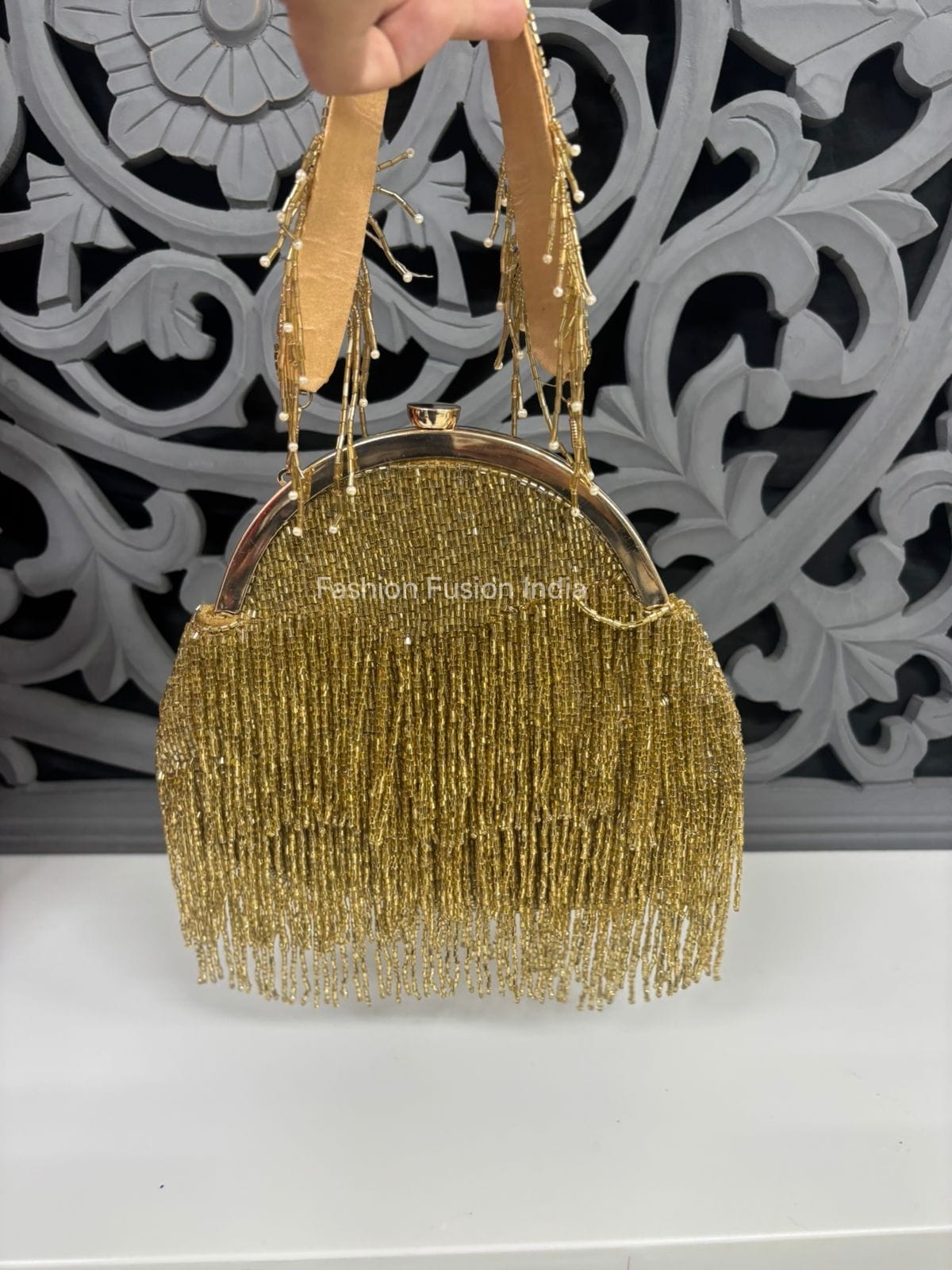 Gold beaded handbag held by a hand against a decorative wall.