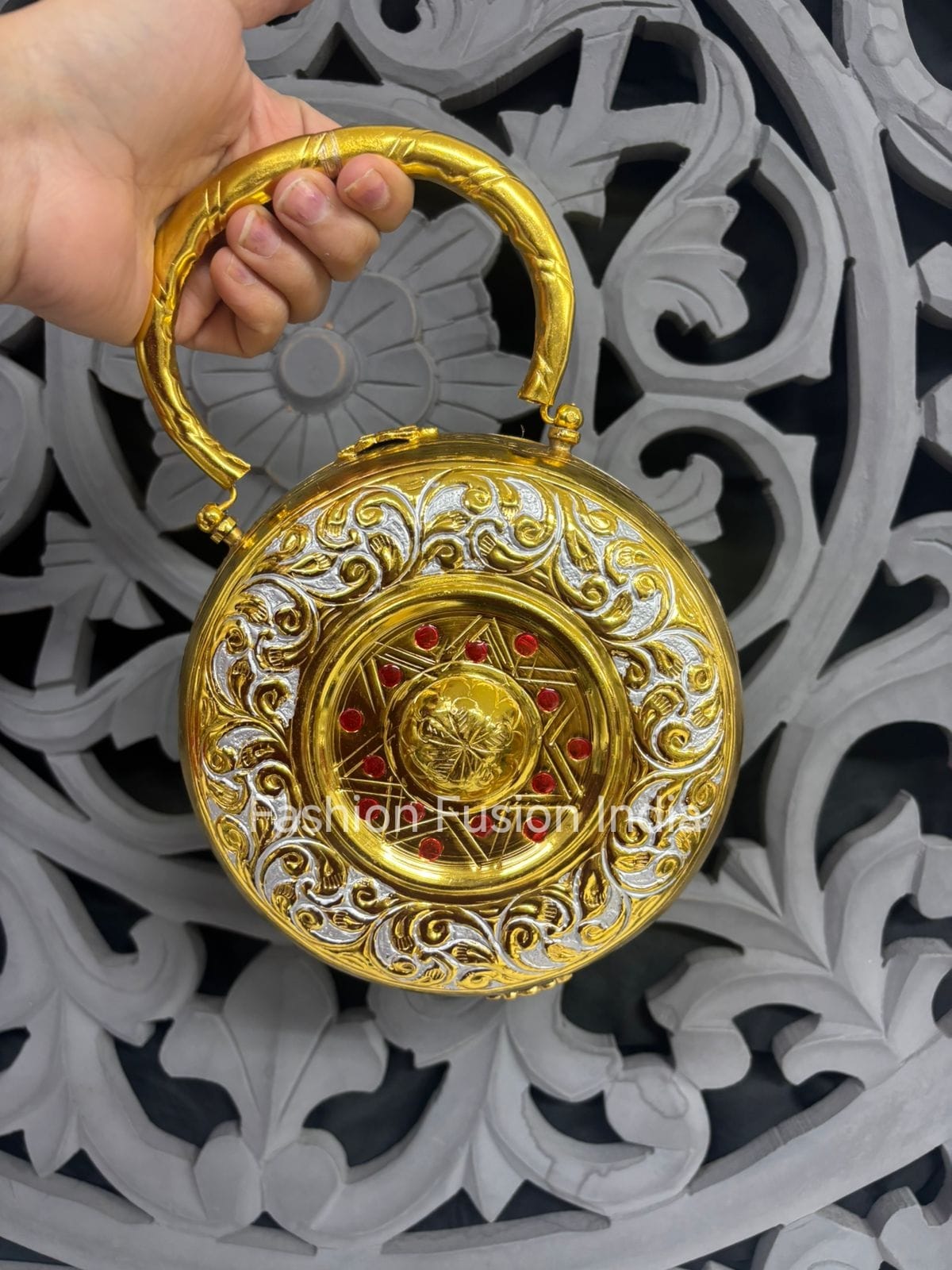 Decorative gold and silver round object with intricate designs held by a hand against an ornate background.
