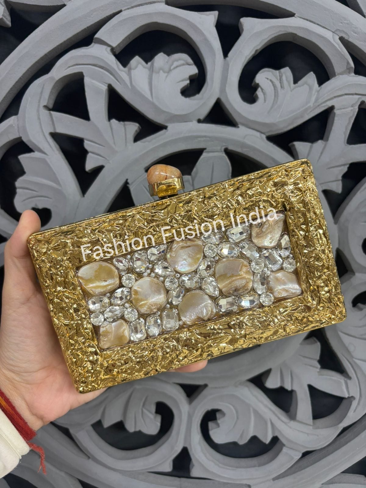 Gold clutch with decorative stones held by a hand against a decorative gray background