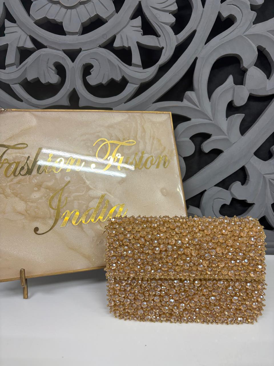 Gold clutch bag in front of a decorative wall with 'Fashion Fusion India' branding.