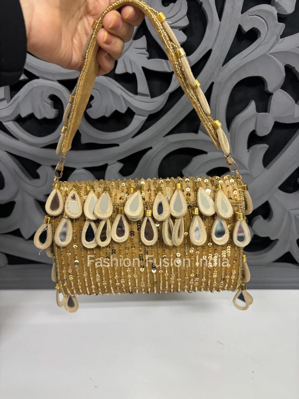 Gold handbag with decorative elements held by a hand against a decorative wall.