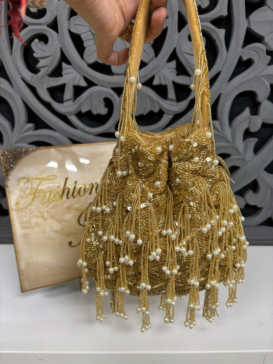 Gold beaded handbag with pearls held by a hand against a decorative wall.