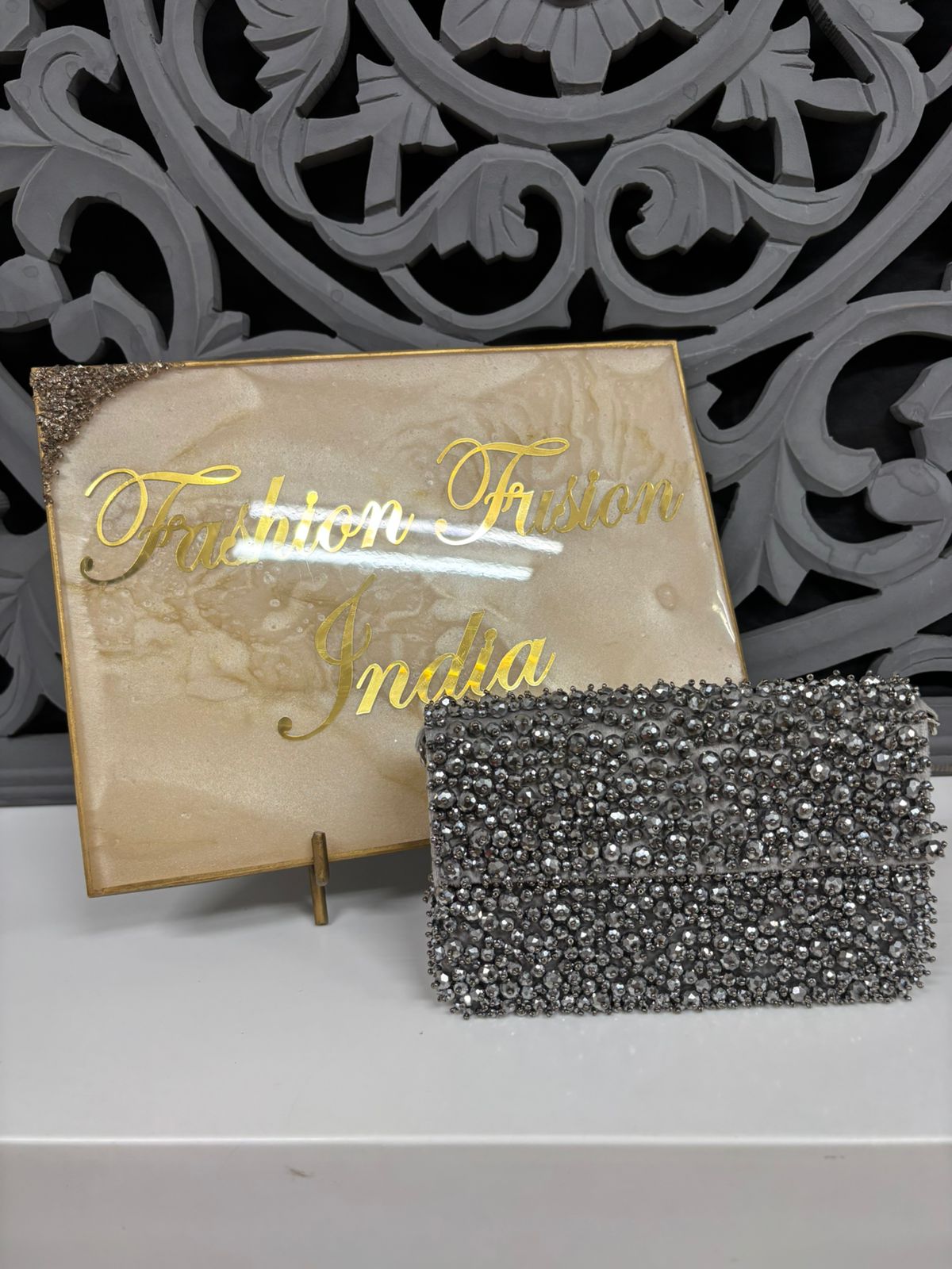 Silver beaded clutch bag in front of a decorative wall with 'Tribhuvan India' branding.