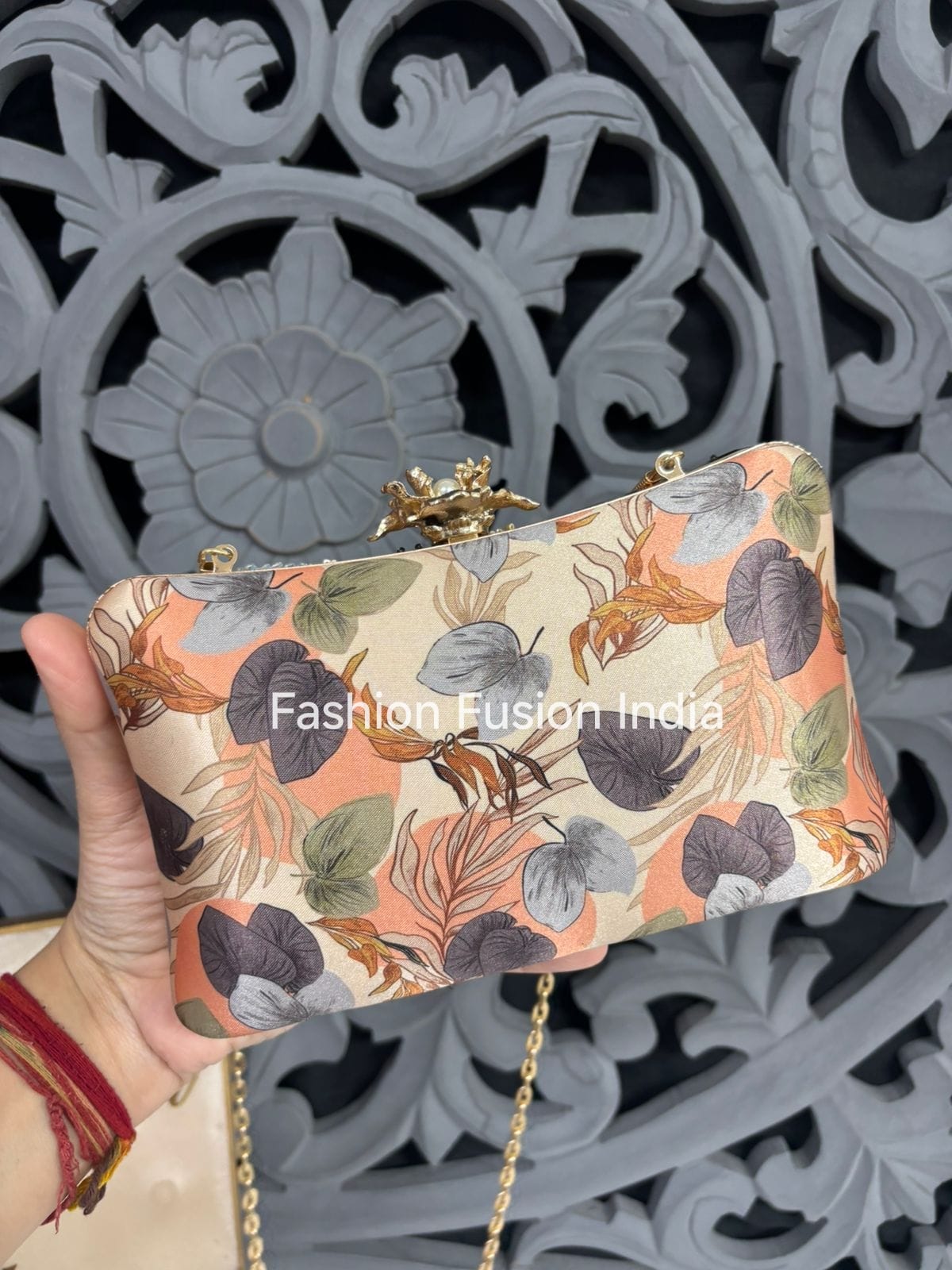 Hand holding a floral clutch bag with 'Fashion Fusion India' branding against an ornate metal background.