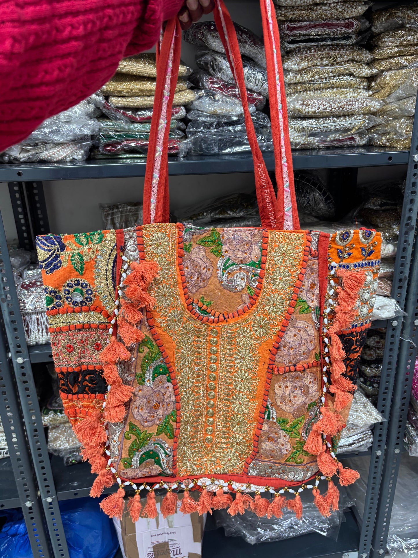 Decorative handbag with intricate patterns and tassels held by a person, with shelves of similar bags in the background.