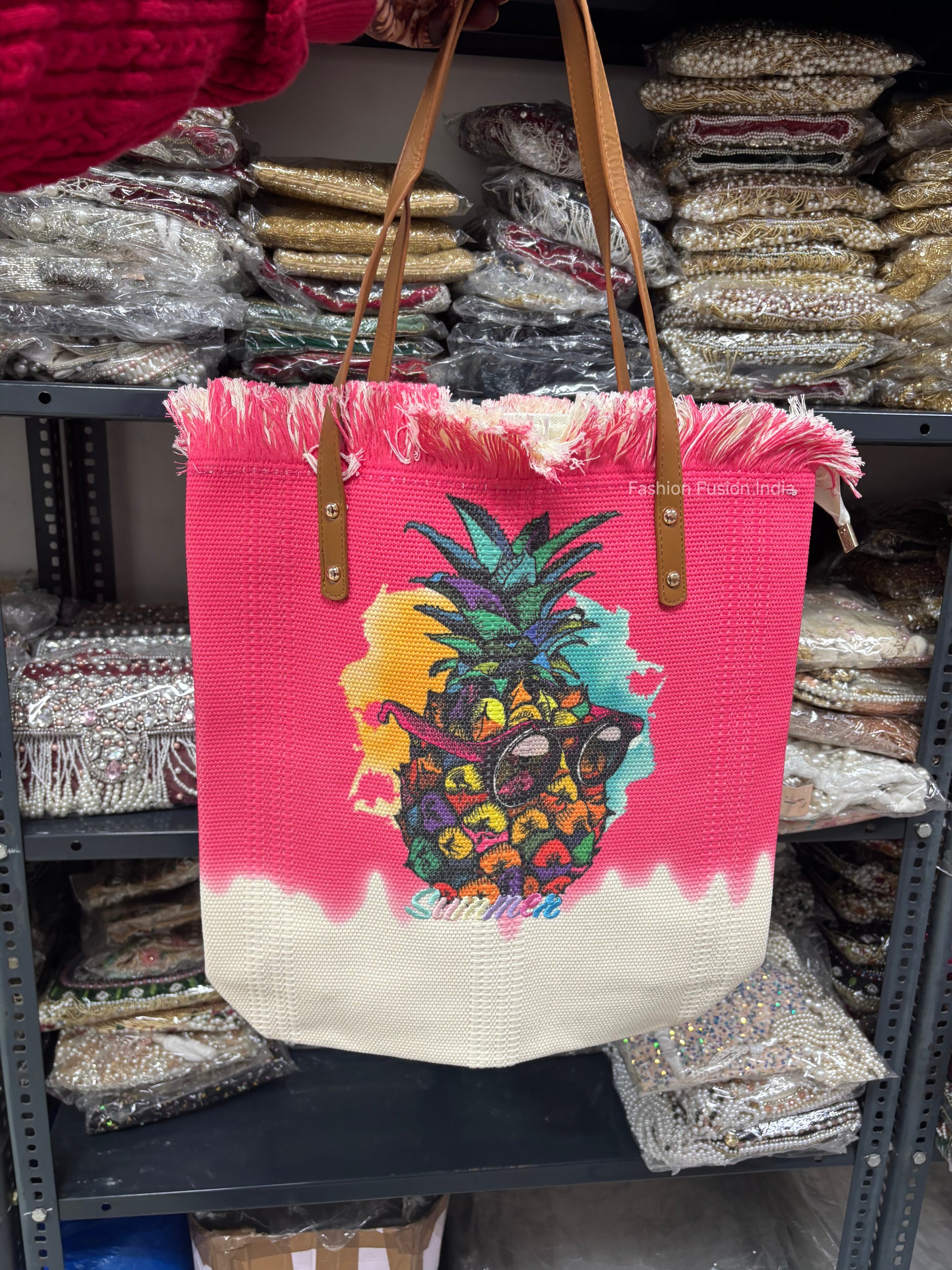 Pink tote bag with colorful pineapple design held in front of shelves with bags.