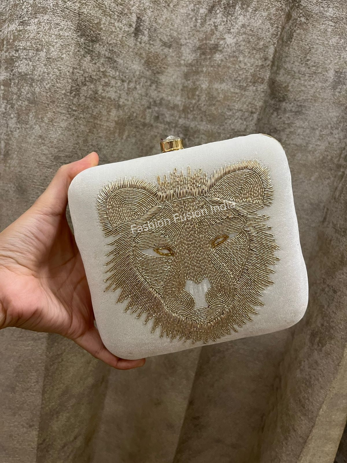 Hand holding a white clutch with gold bear embroidery against a textured beige background