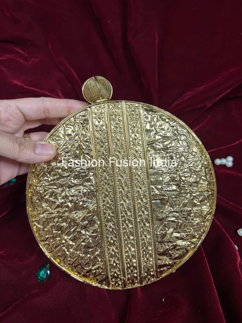 Gold round clutch held by a hand on a red velvet background with 'Fashion Fusion India' branding.