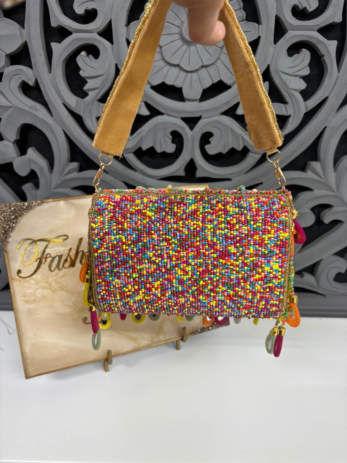 Multicolored beaded handbag with yellow handles on a stand against a decorative wall.