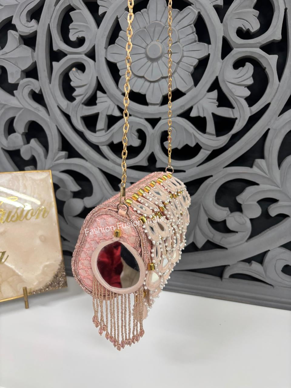 Decorative pink handbag with gold chain and tassels against a decorative gray background.
