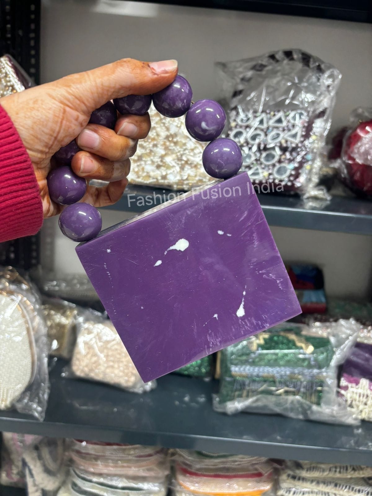 Hand holding a purple bar of soap with beads, against a blurred background of shelves.