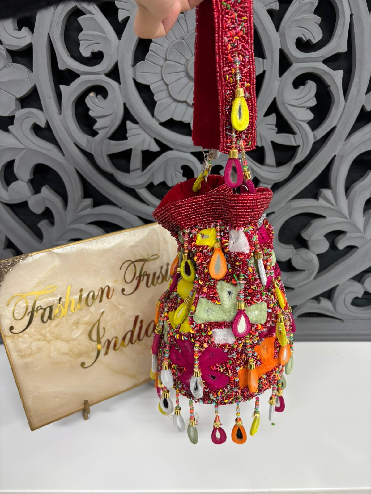Decorative bag with colorful beads and charms held by a hand, with 'Fashion First India' sign in the background.