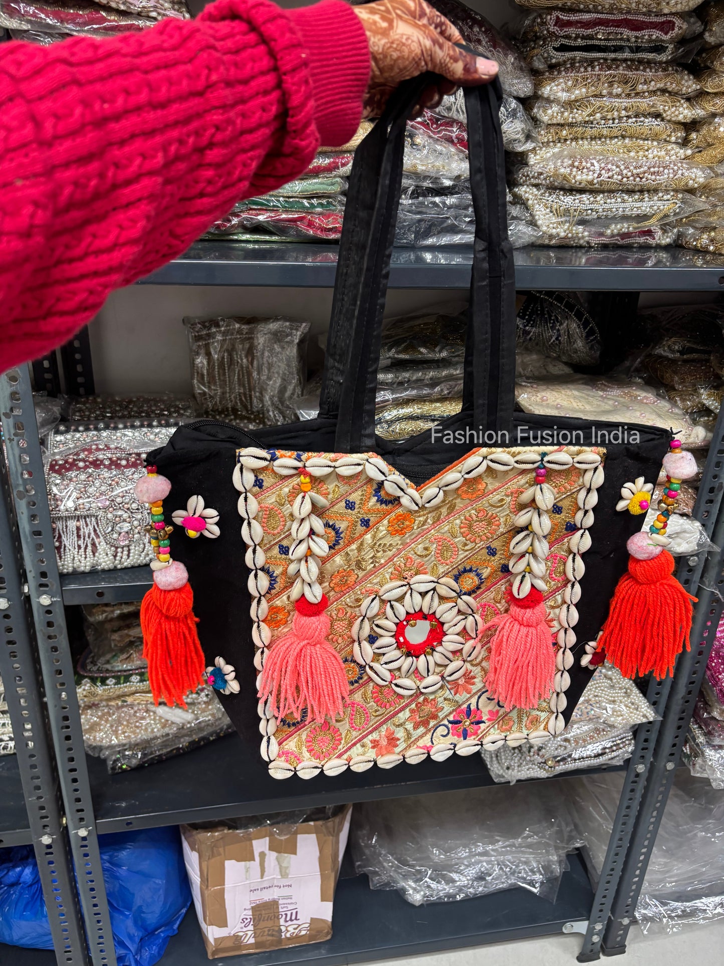 Handbag with decorative elements held by a person in a pink sweater, with a blurred background of shelves.