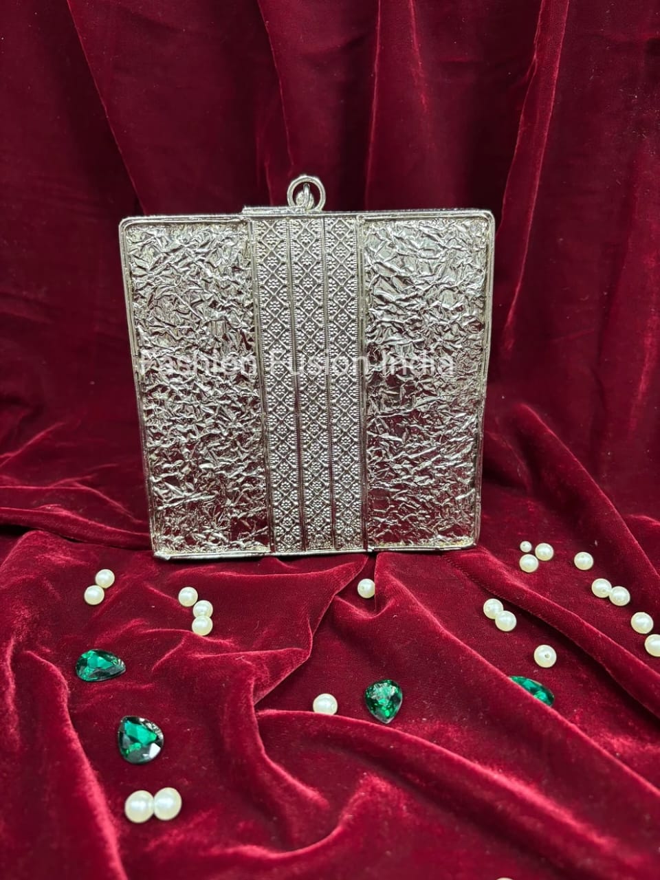Silver clutch bag on a red fabric background with pearls and green stones.