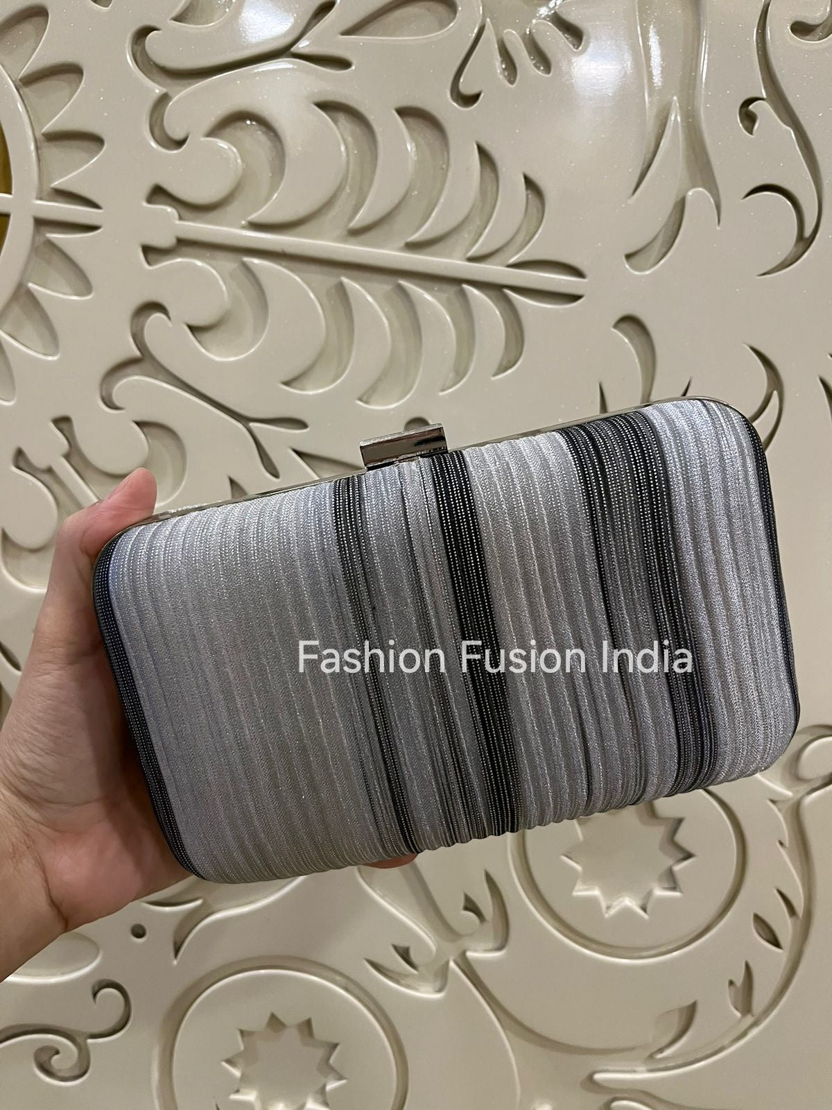 Hand holding a striped clutch bag against an ornate beige background with 'Fashion Fusion India' branding.